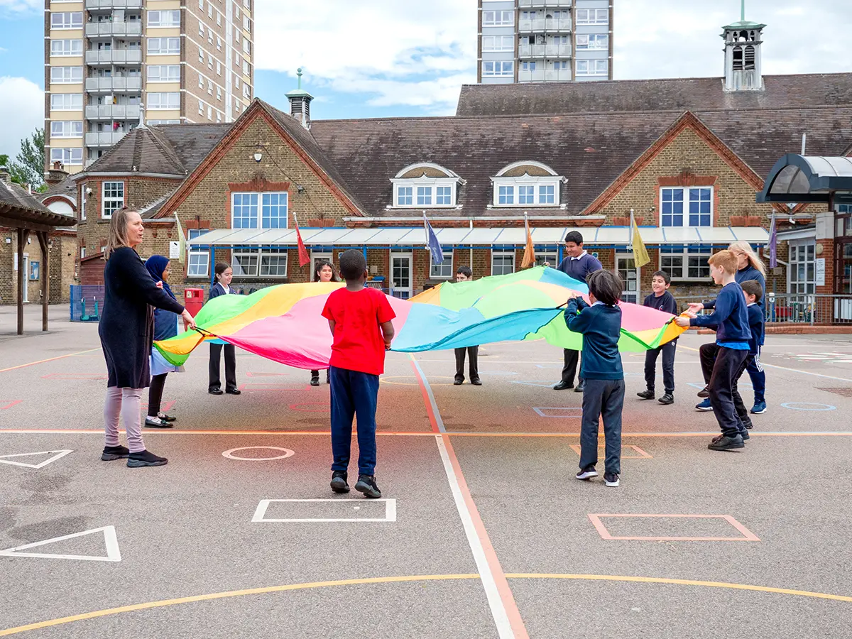 Children and teacher playing with rainbow parachute in the playground
